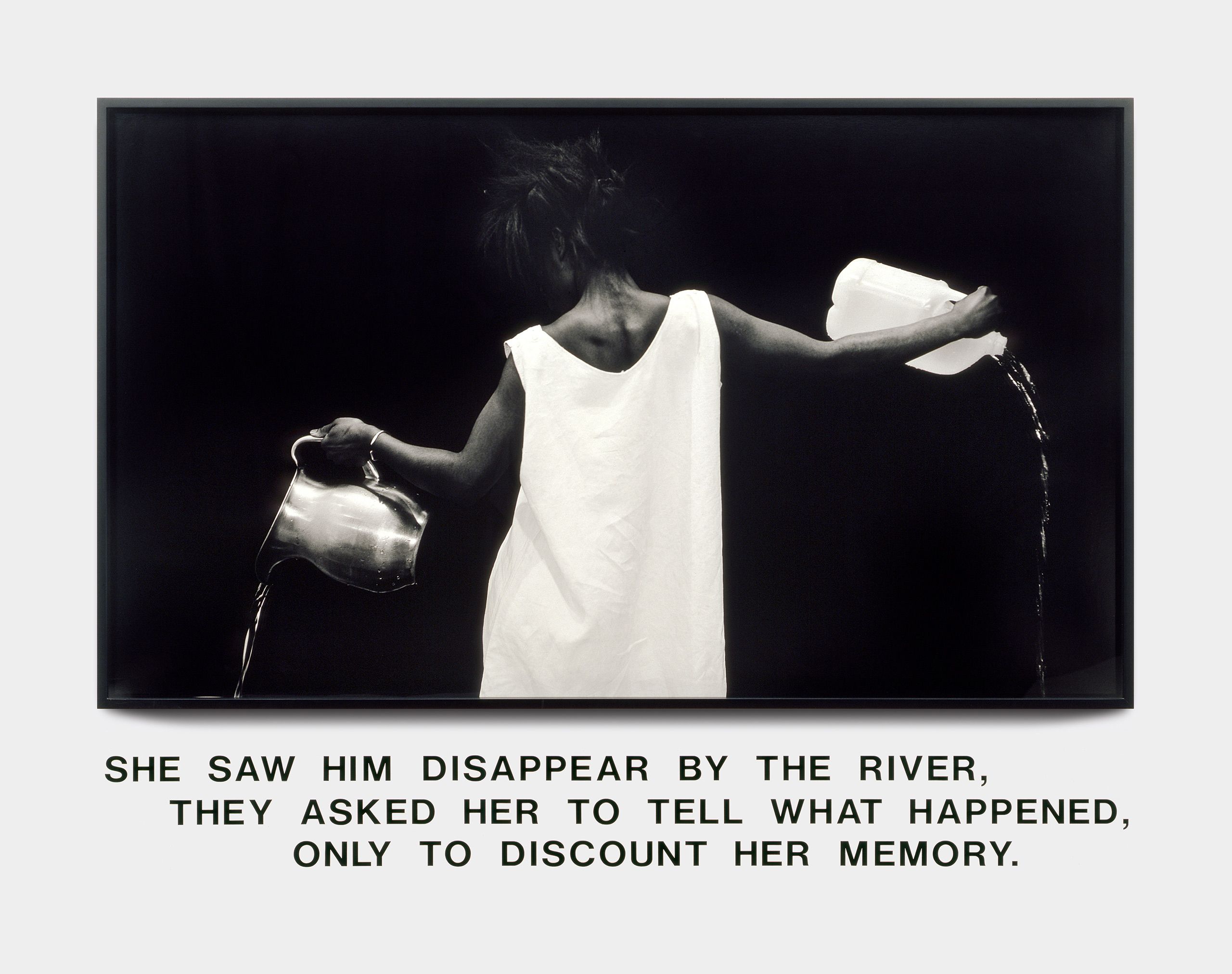 Lorna Simpson's Waterbearer (1986) is a photograph of a Black woman wearing a white shift dress. With her back to the camera, she pours water from two different vessels—a silver pitcher in her left hand and a plastic gallon bottle from her right. Below the image, a text reads, "She saw him disappear by the river, they asked her to tell what happened, only to discount her memory."