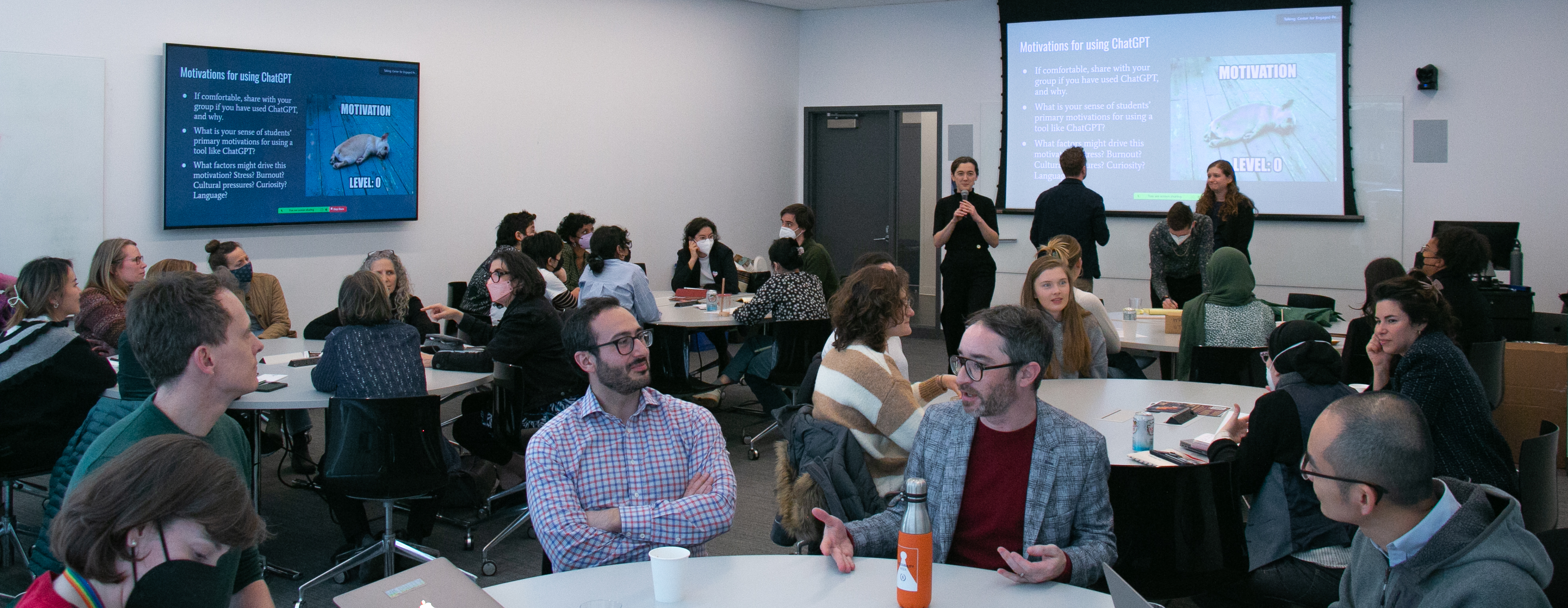 Classroom with several full round tables and facilitators standing. Screens display a meme about low motivation.