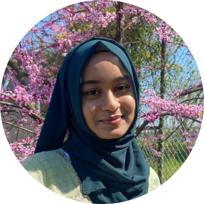 Nayma smiles at the camera with her mouth closed, wears a hijab, and stands in front of cherry blossom trees.