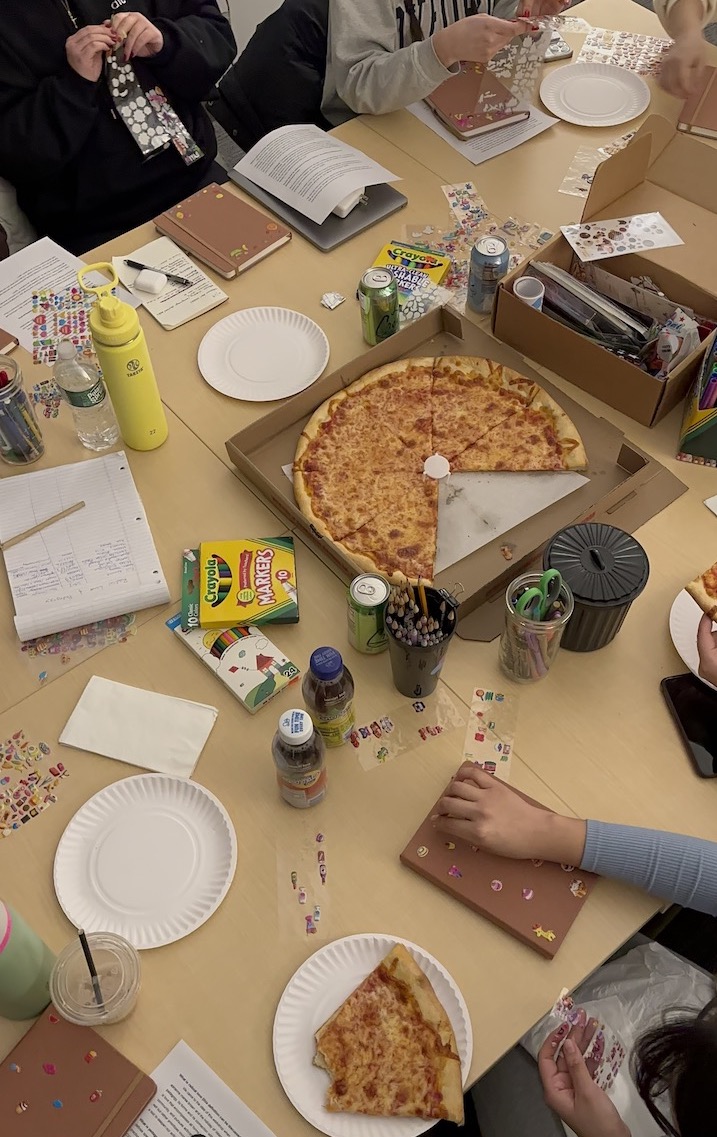A partially eaten pizza sits on a table. There are notebooks, art supplies, writing utensils and drinks on the table. In the lower right corner an arm reaches to put a sticker on a journal.
