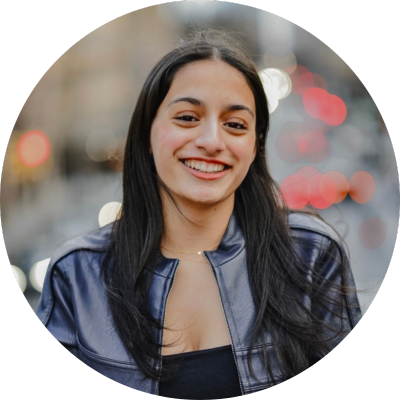 Mira has long hair and wears a leather jacket. She smiles in front of a blurred city background.