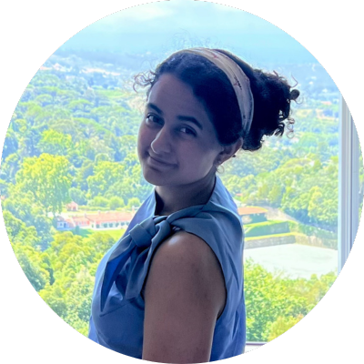 Zala looks over her left shoulder and smiles. She is wearing a headband, and her hair is tied back. There are trees in the background. She is wearing a blue shirt.