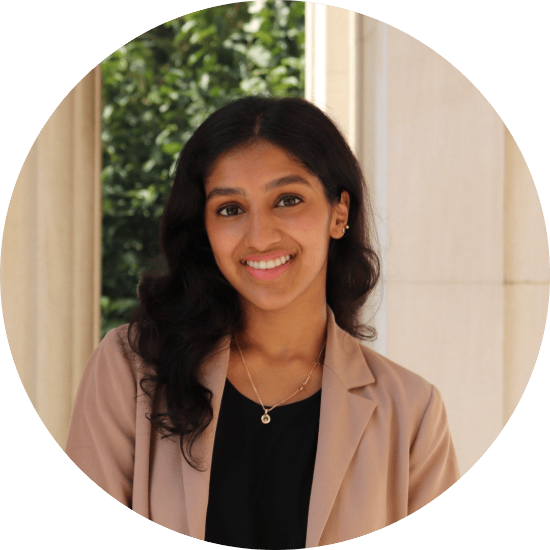 Riya wears a blazer and smiles in front of a pillar.