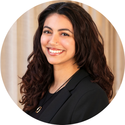 Emma wears a black blazer and a gold pendant necklace. She has curly hair and smiles at the camera.