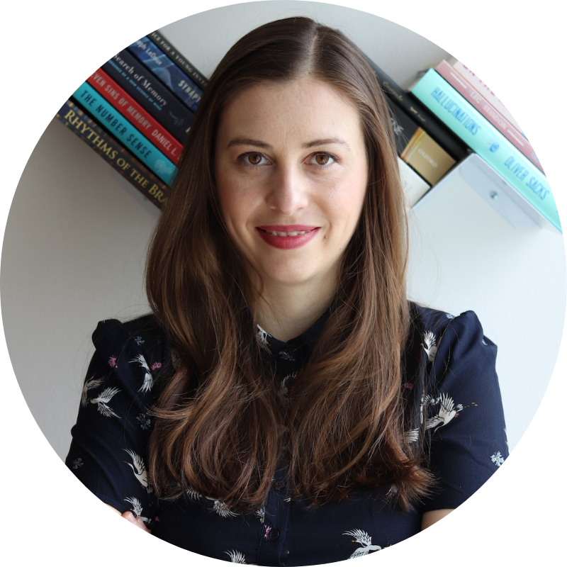 Anamaria Alexandrescu stands in front of a bookshelf and smiles. She is wearing lipstick and a dark blue top. She has long hair.