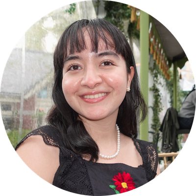 Camila wears a black dress with a red flower on it. She has bangs and smiles at the camera with her mouth open.