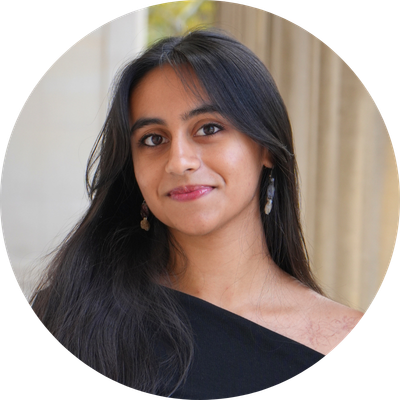 Divya has straight dark hair and smiles with her mouth closed. She stands in front of a pillar.
