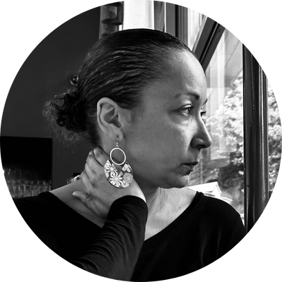 Jennifer Christine Nash rests her hand on her neck and gazes out of a window to the right with her mouth closed. She wears flowery hoop earrings, and her curly hair is tied back in a low bun. She wears a black shirt. 