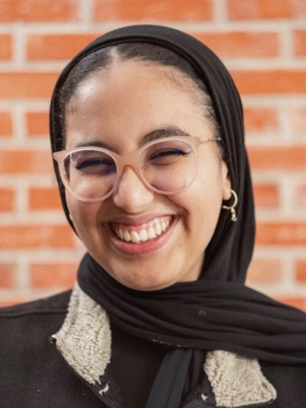 Rayhana wears a black hijab and pink glasses while smiling at the camera. She is in front of a brick wall.