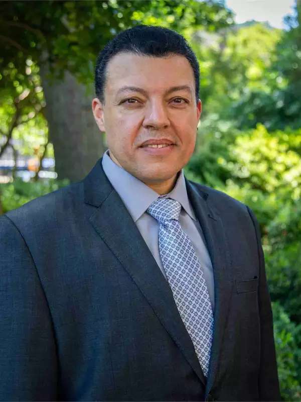 Ahmed Ibrahim wears a suit and smiles in front of the greenery on Barnard's campus. 