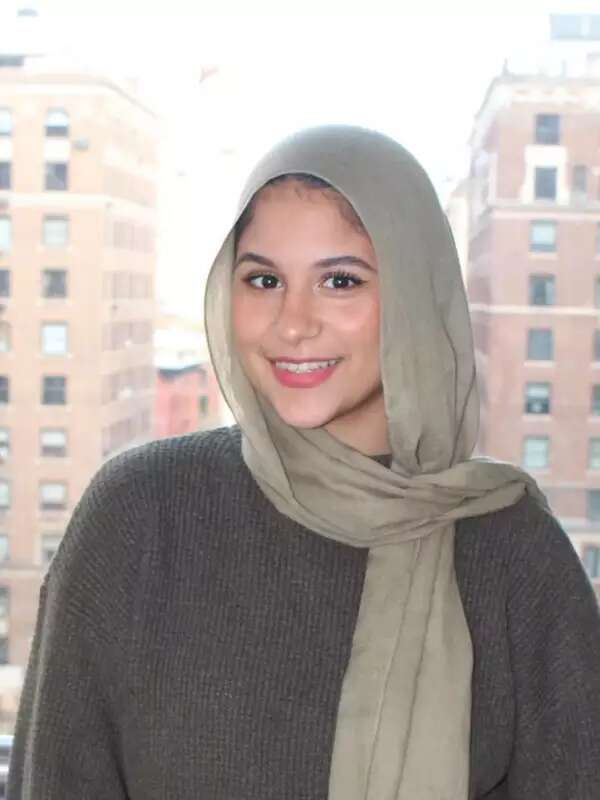 Nada wears hijab and a gray, long-sleeved shirt. She is smiling with her mouth open in front of a window.