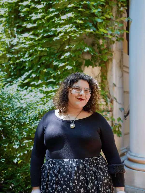 Sydney Smiles in front of an ivy-covered building. They are wearing a black shirt and a patterned skirt.