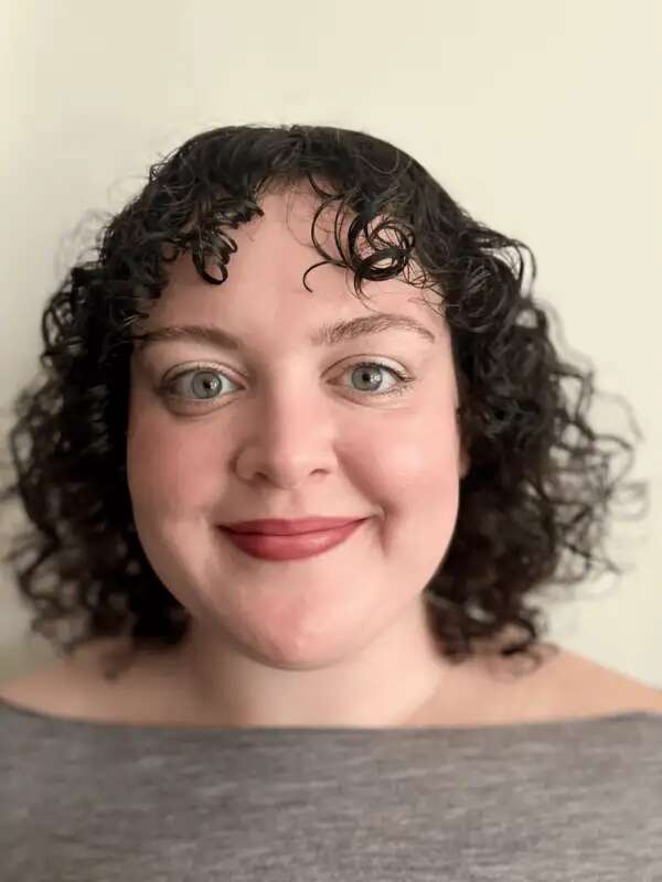 Maddie, a white woman with shoulder length curly hair, looks into the camera and smiles with her mouth closed.