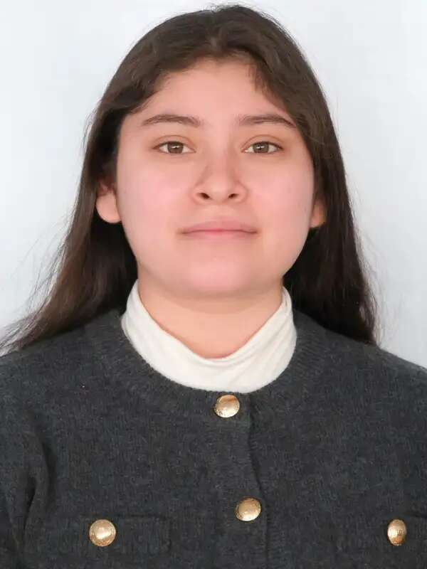 Dayanara looks at the camera and smiles with her chin up and her mouth closed. She is wearing a white turtleneck and a gray light jacket with gold buttons.