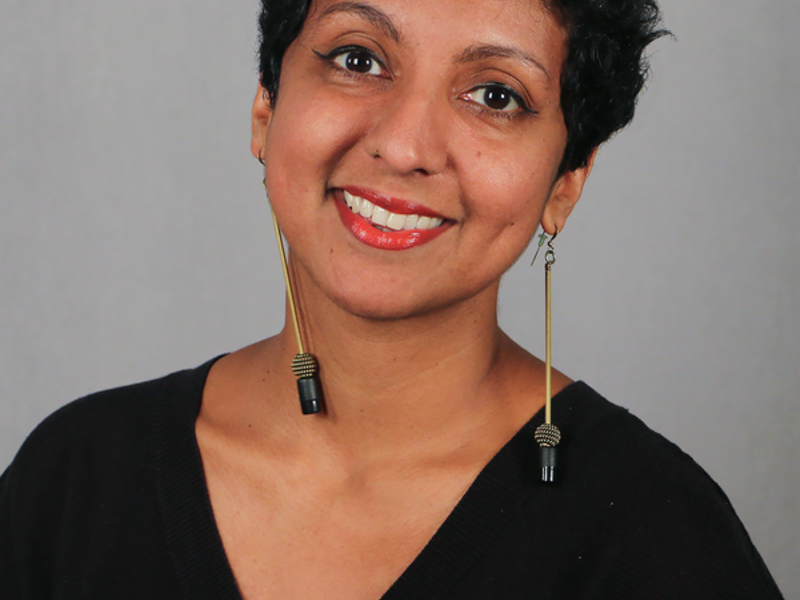 A South Asian person with short black hair wearing a black V-neck smiles into the camera.