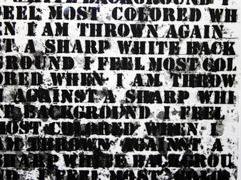 A detail from Glenn Ligon's Untitled (I Feel Most Colored...) (1992), a text-based work that features the phrase "I feel most colored when I am thrown against a sharp white background" screen-printed multiple times over a canvas. As the text repeats, the ink becomes heavier, blacking out the page until the text becomes illegible.