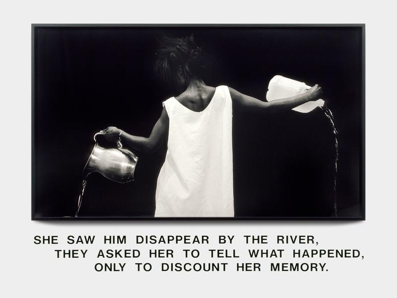 Lorna Simpson's Waterbearer (1986) is a photograph of a Black woman wearing a white shift dress. With her back to the camera, she pours water from two different vessels—a silver pitcher in her left hand and a plastic gallon bottle from her right. Below the image, a text reads, "She saw him disappear by the river, they asked her to tell what happened, only to discount her memory."